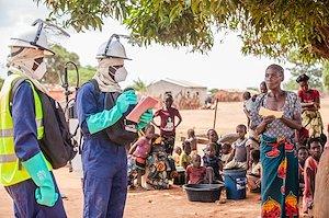 Two men wearing protective clothing speaking with a group of community members under a tree.