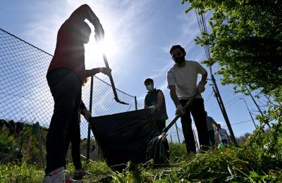 Three participants of a community clean-up action in Kumanovo, North Macedonia shovel waste into a wheelbarrow, backlit by the bright sun.