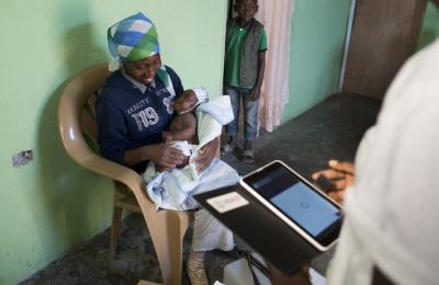A healthcare worker holds tablet at a home health care visit, while a mother and her baby are seated in a chair. 