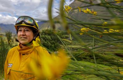 A smiling woman in yellow firefighter gear stands in a green valley surrounded by tall mountains
