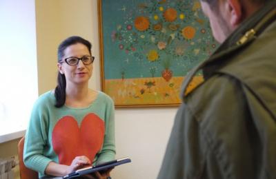 A woman wearing glasses and a green sweater with a large red heart speaking to a man in a room with a floral painting.