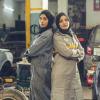 Two female auto shop workers wearing hijabs stand back-to-back in a shop garage.