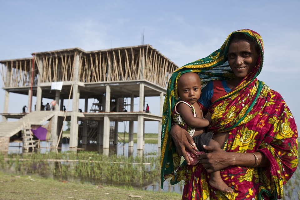 A woman holds her infant in front of a house. Photo credit: Abir Abdullah